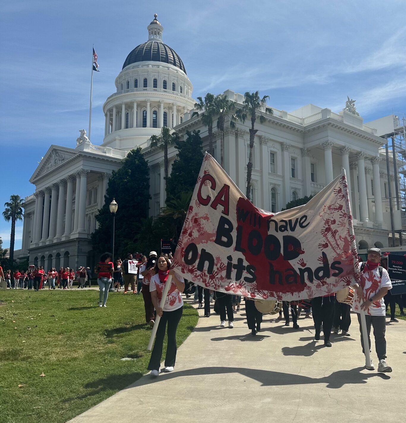 Photo of rally crown in front of the California Capitol marching towards the Swing Space. The large poster on the bottom center-right of the photo states "CA will have blood on its hands" and is covered with red handprints and a red colored in drawing of California state.